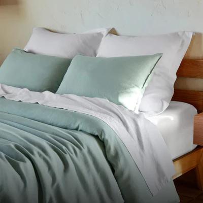 Bed made with green and white bedding and pillows in a softly lit room with a white textured wall.